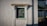 A small rectangular window set in a textured white wall. The window has a green frame and is topped with a decorative wooden canopy. A black drainpipe runs vertically next to the window. To the right, there is a shaded area with a green roof supported by metal framework.