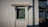 A small rectangular window set in a textured white wall. The window has a green frame and is topped with a decorative wooden canopy. A black drainpipe runs vertically next to the window. To the right, there is a shaded area with a green roof supported by metal framework.