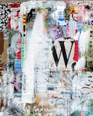 An abstract collage featuring layers of various textures, printed text, and colors. Elements include torn paper with fragments of text and images, paint splatters, and brush strokes. A large letter 'W' and a mix of vibrant colors with white, black, and earth tones are prominent.