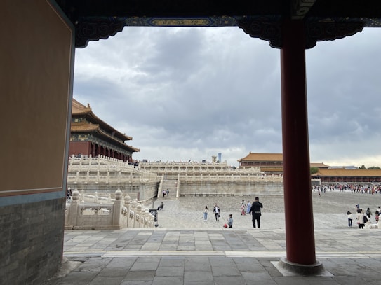A view of an ancient architectural complex with intricate stone carvings and a grand structure with a traditional Chinese roof. The area is populated with visitors walking and exploring the site, under a cloudy sky that sets a moody atmosphere.