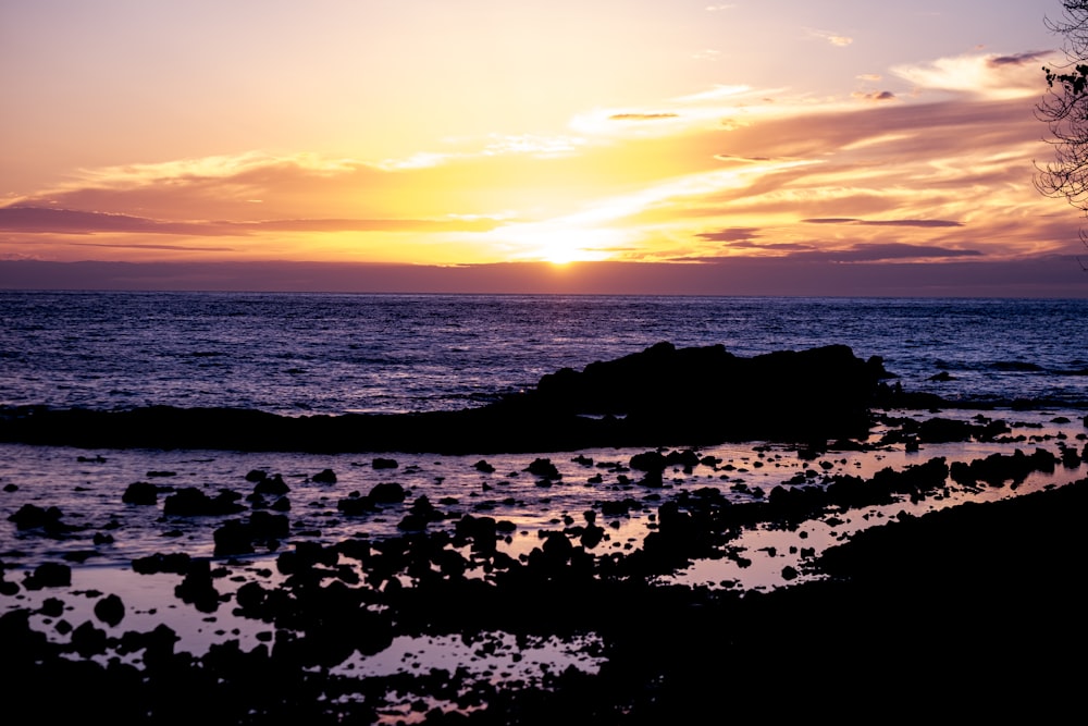 the sun is setting over the ocean with rocks in the foreground