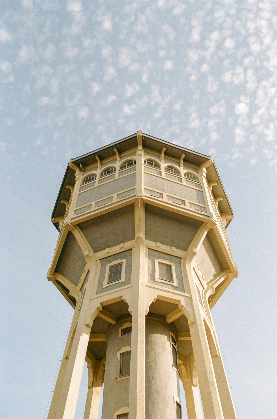 The image shows a tall, cylindrical water tower with a pointed roof and multiple levels. The tower is painted white and stands against a clear blue sky dotted with fluffy white clouds. The perspective of the photo is from below, looking up at the imposing structure.