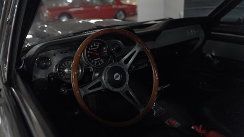 A classic car interior with a wooden steering wheel is visible, featuring a prominent tachometer and various gauges on the dashboard. The dim lighting gives a vintage ambiance, while the reflections on the windshield point to cars parked outside.