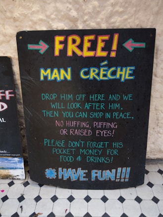 A humorous chalkboard sign titled 'FREE! MAN CRÉCHE' offers to look after men while their companions shop in peace. It humorously suggests not to forget their pocket money for food and drinks and ends with 'HAVE FUN!!!' in colorful lettering.