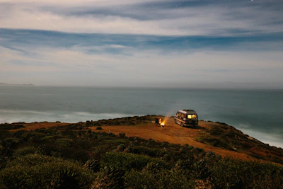 A lone male traveler setting up camp next to his van on a coastal cliff at sunset.