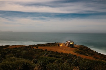 A scenic coastal view with a vintage van parked on a grassy cliff overlooking the ocean. A campfire is lit beside the van, casting a warm glow. The sky is vast and mostly clear, with a few clouds adding texture.