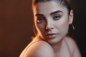 A close-up portrait of a woman with elegant makeup and subtle lighting. Her hair is neatly tied back, and she is wearing large hoop earrings. The lighting creates a soft, warm glow, accentuating her features.