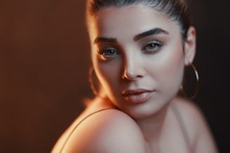A close-up portrait of a woman with elegant makeup and subtle lighting. Her hair is neatly tied back, and she is wearing large hoop earrings. The lighting creates a soft, warm glow, accentuating her features.