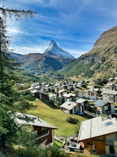 A picturesque mountain village nestled among verdant hills, with traditional wooden houses scattered across the landscape. In the background, a stunning snow-capped peak rises majestically against a clear blue sky, surrounded by rugged mountain terrain.
