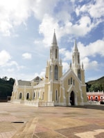 A large, ornate church with two tall spires, featuring arched windows and intricate architectural details. The building is painted in a light beige color with white accents, surrounded by a spacious paved area. In the background, there are green trees and a hillside. Bright blue sky with scattered clouds completes the serene setting.