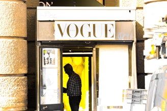 A person in a checkered shirt is seen in silhouette standing at the entrance of a store with a sign that reads 'VOGUE'. The background inside the store is brightly lit with yellow lighting and what appears to be yellow balloons. The store’s exterior has a rough stone wall on both sides of the entrance.