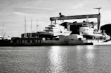 A waterfront shipyard featuring several large yachts docked alongside various structures, including a prominent crane labeled 'LA CIOTAT 105.' The water in the foreground reflects light, adding a serene ambiance. The background contains a mix of industrial elements and a mountainous landscape.
