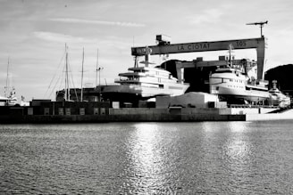 A waterfront shipyard featuring several large yachts docked alongside various structures, including a prominent crane labeled 'LA CIOTAT 105.' The water in the foreground reflects light, adding a serene ambiance. The background contains a mix of industrial elements and a mountainous landscape.