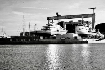 A waterfront shipyard featuring several large yachts docked alongside various structures, including a prominent crane labeled 'LA CIOTAT 105.' The water in the foreground reflects light, adding a serene ambiance. The background contains a mix of industrial elements and a mountainous landscape.