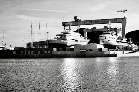 A waterfront shipyard featuring several large yachts docked alongside various structures, including a prominent crane labeled 'LA CIOTAT 105.' The water in the foreground reflects light, adding a serene ambiance. The background contains a mix of industrial elements and a mountainous landscape.