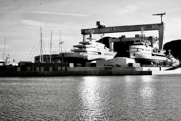 A waterfront shipyard featuring several large yachts docked alongside various structures, including a prominent crane labeled 'LA CIOTAT 105.' The water in the foreground reflects light, adding a serene ambiance. The background contains a mix of industrial elements and a mountainous landscape.