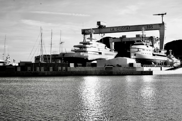 A waterfront shipyard featuring several large yachts docked alongside various structures, including a prominent crane labeled 'LA CIOTAT 105.' The water in the foreground reflects light, adding a serene ambiance. The background contains a mix of industrial elements and a mountainous landscape.