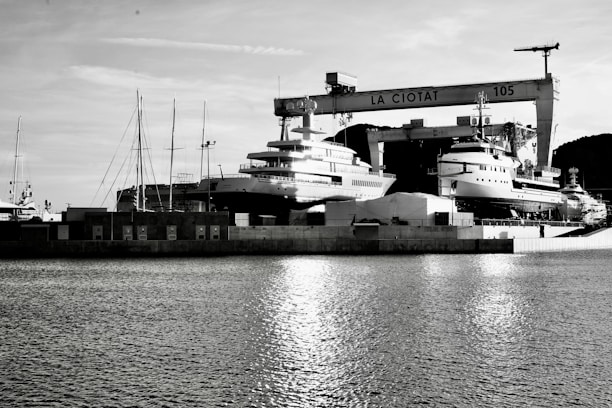 A waterfront shipyard featuring several large yachts docked alongside various structures, including a prominent crane labeled 'LA CIOTAT 105.' The water in the foreground reflects light, adding a serene ambiance. The background contains a mix of industrial elements and a mountainous landscape.