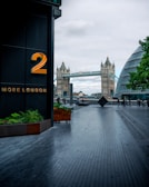 A modern urban scene featuring a walkway with dark paving stones leading towards the iconic Tower Bridge in the background. On the left, a building with a large number '2' and the words 'MORE LONDON' in gold lettering. On the right, there is a modern glass building with a curved design. Several people are walking along the path, and planters with green plants are visible.