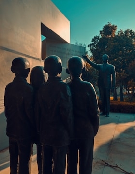 A group of statues depicting four children stand facing a stone structure with engravings. In the distance, there is an adult figure with an outstretched hand, seemingly guiding or teaching the children. The scene is set outdoors, with trees and a paved pathway visible.