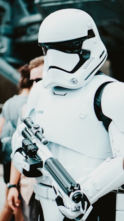 A person wearing a futuristic white armor suit, equipped with a helmet that covers the entire face and a chest plate. The attire is sleek, with black accents and a prominent visor on the helmet. In the hands, there is a detailed and sophisticated weapon, appearing to be a blaster. Background elements include blurred individuals, possibly indicating a public setting.
