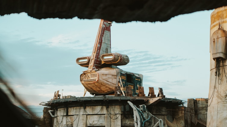 A futuristic spacecraft is perched on a circular platform amid a desert environment. The spacecraft has a weathered appearance, with a mix of beige and red colors. The sky is overcast with scattered clouds, creating a serene atmosphere.