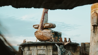 A futuristic spacecraft is perched on a circular platform amid a desert environment. The spacecraft has a weathered appearance, with a mix of beige and red colors. The sky is overcast with scattered clouds, creating a serene atmosphere.