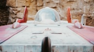 A futuristic-looking spacecraft with a sleek, aerodynamic shape is positioned against a textured stone wall. The vehicle features a glass cockpit and red accents, giving it a vintage sci-fi aesthetic.