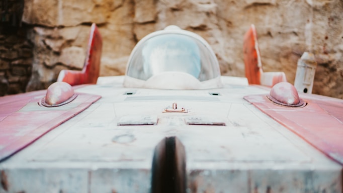 A futuristic-looking spacecraft with a sleek, aerodynamic shape is positioned against a textured stone wall. The vehicle features a glass cockpit and red accents, giving it a vintage sci-fi aesthetic.