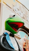 A large statue of a green frog wearing a tuxedo with a bow tie is prominently displayed. The background features architectural details and an image of a pig character.