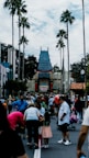 A crowded theme park entrance with many people gathered around. In the background, a distinctive Chinese-style building with ornate roof detailing stands prominently. Tall palm trees line the pathway, adding a California-influenced atmosphere.