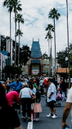 A crowded theme park entrance with many people gathered around. In the background, a distinctive Chinese-style building with ornate roof detailing stands prominently. Tall palm trees line the pathway, adding a California-influenced atmosphere.