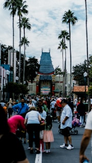 A crowded theme park entrance with many people gathered around. In the background, a distinctive Chinese-style building with ornate roof detailing stands prominently. Tall palm trees line the pathway, adding a California-influenced atmosphere.