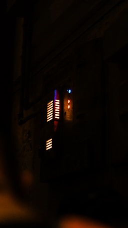 A dimly lit control panel or display board with illuminated colored lights including red, blue, and yellow glowing indicators against a dark background. The setting suggests a mysterious or industrial environment.
