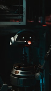 A robot with a cylindrical body and a round head featuring mechanical eyes and a red light in the center. The robot includes metallic parts and appears to be in a dimly lit industrial setting with metal bars in the background.