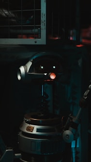 A robot with a cylindrical body and a round head featuring mechanical eyes and a red light in the center. The robot includes metallic parts and appears to be in a dimly lit industrial setting with metal bars in the background.