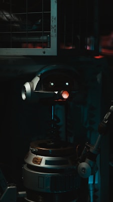 A robot with a cylindrical body and a round head featuring mechanical eyes and a red light in the center. The robot includes metallic parts and appears to be in a dimly lit industrial setting with metal bars in the background.