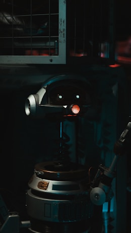 A robot with a cylindrical body and a round head featuring mechanical eyes and a red light in the center. The robot includes metallic parts and appears to be in a dimly lit industrial setting with metal bars in the background.