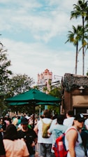 A bustling outdoor scene in a theme park with a prominent building resembling the Hollywood Tower Hotel in the background. The area is crowded with people wearing casual summer clothing, some under a large green umbrella. Palm trees and lush greenery add to the lively atmosphere, with the sky partially cloudy.