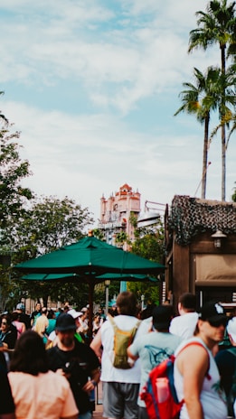 A bustling outdoor scene in a theme park with a prominent building resembling the Hollywood Tower Hotel in the background. The area is crowded with people wearing casual summer clothing, some under a large green umbrella. Palm trees and lush greenery add to the lively atmosphere, with the sky partially cloudy.