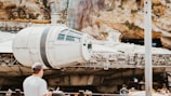 A large, detailed spacecraft displayed outdoors against a rocky and textured backdrop, with several people standing nearby observing. The atmosphere suggests a science fiction or futuristic theme park setting.