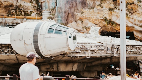 A large, detailed spacecraft displayed outdoors against a rocky and textured backdrop, with several people standing nearby observing. The atmosphere suggests a science fiction or futuristic theme park setting.