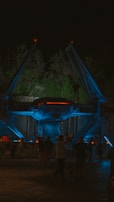 A futuristic spacecraft is displayed at night under atmospheric lighting with blue and red accents. The craft is surrounded by trees, and several people are walking around, suggesting an outdoor exhibition or theme park setting.