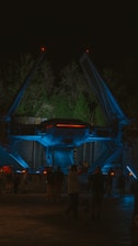 A futuristic spacecraft is displayed at night under atmospheric lighting with blue and red accents. The craft is surrounded by trees, and several people are walking around, suggesting an outdoor exhibition or theme park setting.