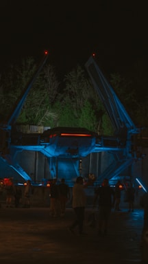 A futuristic spacecraft is displayed at night under atmospheric lighting with blue and red accents. The craft is surrounded by trees, and several people are walking around, suggesting an outdoor exhibition or theme park setting.