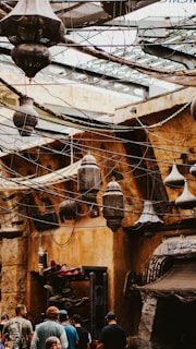 A bustling market area with a rustic, old-world charm featuring hanging lanterns and an intricate network of wires above. The walls have a warm, earthy tone, and elements of traditional design are evident. People are gathered, walking or standing in groups, suggesting a lively and engaging environment.