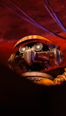 A robot with intricate details is positioned against a dimly lit, reddish background. The robot features a pair of goggles with round lenses and a grill-like mouth. Orange accents are present on its headgear, resembling a cap or visor. The metallic texture and design components suggest a blend of retro and industrial aesthetics.