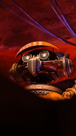 A robot with intricate details is positioned against a dimly lit, reddish background. The robot features a pair of goggles with round lenses and a grill-like mouth. Orange accents are present on its headgear, resembling a cap or visor. The metallic texture and design components suggest a blend of retro and industrial aesthetics.