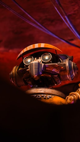 A robot with intricate details is positioned against a dimly lit, reddish background. The robot features a pair of goggles with round lenses and a grill-like mouth. Orange accents are present on its headgear, resembling a cap or visor. The metallic texture and design components suggest a blend of retro and industrial aesthetics.