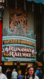 A theme park attraction entrance featuring a vibrant sign with characters and colorful lights. The sign reads 'Mickey & Minnie's Runaway Railway' and there are people walking beneath it. Above the sign, an intricate dragon design is incorporated into the structure.